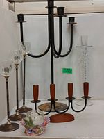 Front view of assorted candlesticks showing large black metal four-arm piece, glass twisted column candlestick, tall glass candlesticks with metal bases, and small floral decorative glass holder.
