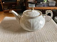 Side view of white porcelain teapot showing embossed petal design and gold accents on spout tip, lid knob, and handle.