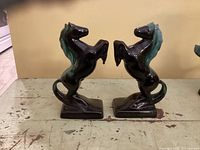 Pair of glossy black rearing horse bookends with greenish-blue manes standing on rectangular bases, placed on a worn wooden surface with a beige background.