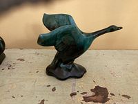 Blue and green ceramic goose figurine standing with wings raised on a dark base.
