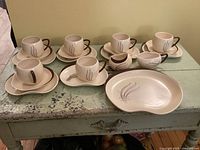 Six cream cups with brown handles, five saucers, six small plates, cream and sugar set and a serving plate, all on a rustic table.