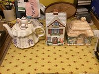 Photo showing ceramic teapot in dress shape with purple bows, bird lid, and two ceramic house canisters
