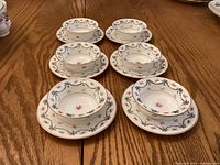 Full view of six small bowls each set on a matching saucer, white porcelain with pink rose and green ribbon pattern, gold rims.