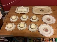 Full set layout on wooden table showing two plates, six cups with saucers, and rectangular serving platter with covered dish at upper left