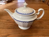 Side view of white porcelain teapot with blue floral trim and gold accents on handle and spout, sitting on wood surface.