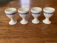 Four white porcelain Hutschenreuther egg cups with blue decoration around rim, arranged side by side on wooden table.