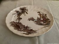 Front view of the oval platter showing The Old Mill scene with trees, water, and buildings in brown transferware design on cream ceramic with scalloped edges.