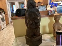 Front view of large carved wooden sculpture of a pregnant nude woman, showing prominent breasts and belly, placed on a kitchen counter.