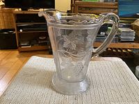 Clear pressed glass pitcher with grape and vine pattern viewed from front showing body, handle, and spout.