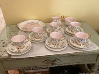 Full set arranged on a distressed wooden surface showing cups on saucers, bread plates behind, and creamer, sugar bowl, and cake plate in back