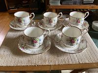 Five bone china teacups and saucers displayed on a woven table mat, showing the floral design and gold trim.
