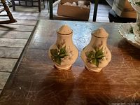 Pair of ceramic salt and pepper shakers with green leaf and white flower pattern on wooden table.