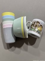 Photo shows six pastel-colored plastic cups, three plastic bowls with pastel rims, and one ceramic mug with a hedgehog illustration and text.