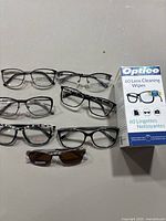 Six eyeglass frames arranged in two rows with a box of Optico lens cleaning wipes on the side.