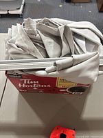 Light beige fabric curtain folded and placed inside a cardboard box with white curtain rods and rail visible on top.