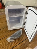 Mini fridge with open door showing interior shelf and vehicle charger wrapped in plastic on wooden floor.