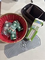 Red woven basket containing multiple small green boxes of Pike Place Nespresso coffee capsules, two brown fabric storage containers, gray rubber sink mat, and open box of wrapped paper straws.