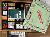 Overhead image showing all components of the multi game set including Monopoly board, Clue instruction booklet, cribbage board, cards, dice, and game tokens arranged inside a wooden storage board with compartments.
