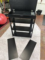 Black wood shoe rack with a top compartment opened showing metal hinges and screws, and three shelves below. Two shelves are placed on the floor in front of the rack.