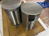 Two stainless steel garbage cans side by side on a wooden table, showing the lids and foot pedals.