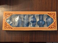 Top view of the set of six blue ceramic bud vases in original box with an orange decorative border and plastic display window