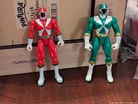 Photo showing standing Red and Green Lightspeed Rescue Power Rangers figures on a wooden surface with cardboard behind.
