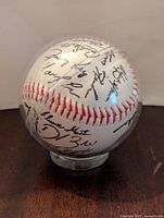 Baseball with black printed facsimile signatures visible on one side in clear display case.