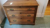 Full frontal view of the wooden antique dresser showing three drawers with brass handles and keyhole escutcheons on a carpeted floor.