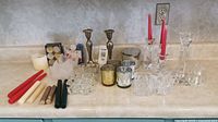 View of assorted glass, metallic votives and candlesticks arranged on a counter with various taper candles and a Bath and Body Works candle.