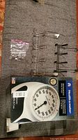 Two metal door hooks stacked side by side with small plastic mounting hardware bag. Health o meter analog bathroom scale in box shown below.