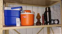Photo showing three coolers, a vintage oil lamp, a stainless steel thermos, and a barometer canteen on a shelf.