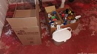 Photo of cleaning supplies in cardboard box, white compost bin and fermenting bottle on red floor.