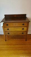 Front view of antique 3-drawer dresser showing the medium-brown drawers, dark wooden top, metal pull handles, and tapered legs.