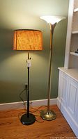 Photo shows two floor lamps side by side against a green wall and hardwood floor. The left lamp has a black round base and brown fabric shade. The right lamp has a gold clover-shaped base and a white upward-facing glass shade.