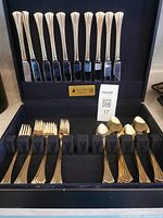 Full set of gold plated flatware displayed in a black presentation case, showing knives in the back and forks and spoons in the front slots.