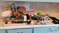 Wide view showing the kitchen counter with wooden utensil block with knives, trays of stainless steel flatware, assorted kitchen utensils in containers, small kitchen tools, and accessories.