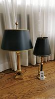 Two gold base table lamps with black shades standing side by side in front of white curtains on hardwood floor.