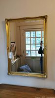 Wall mirror with gold ornate frame reflecting room with door and furniture.