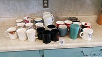 Collection of twenty assorted mugs on countertop including floral, solid colors, white, black, blue, red, teal, and clear glass.