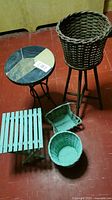 Photo showing one round plant stand with multi-color rock tile top and black metal legs, one tall dark brown wicker basket planter with three wooden legs, one light blue wooden slatted stool, one small rectangular light blue basket, and one round light blue woven basket all placed on a red floor.