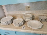 Stacked white octagonal dinner plates, small plates, dessert plates, and bowls set on a kitchen counter.