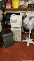 Overview of three dehumidifiers stacked and placed alongside a white standing oscillating fan on the right.