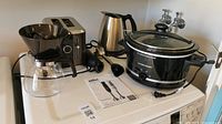 Photo shows the full set of kitchen appliances on a white countertop including crock pot, coffee carafe, toaster, kettle and immersion blender.