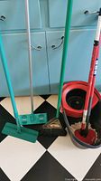 Various kitchen cleaning tools arranged on a black and white checkered floor in front of blue cabinets. Items include floor mop, broom, dustpan, and mop bucket.