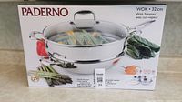 Box front showing Paderno brand, product image of wok with steamer insert and glass lid, and vegetables around it. The box states the wok is 32 cm size and includes a steamer.