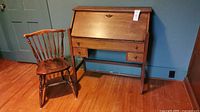 Wooden antique writing desk with slant front door closed, accompanied by matching wooden chair on hardwood floor against blue wall.