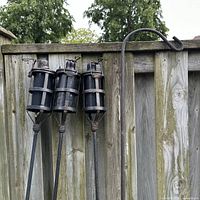 Three metal tiki torches and one wrought iron bird feeder post leaning against a wooden fence outdoors. The torches have a cage-like metal frame surrounding the wick.