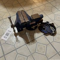 Blue bench vise with 4-inch jaws, visible rust and chipped paint on metal surfaces. Handle installed on side to adjust jaw width. Swivel base detached and sitting next to vise body, appears broken and worn.