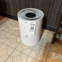 Front view of the white cylindrical Bissell MyAir air purifier with power cord plugged in, showing top vent and logo.