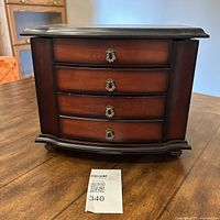Closed wooden jewelry box showing four curved drawers with metal handles on front and classic style base